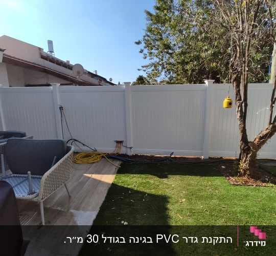 גינה עם דשא סינתטי, עץ, גדר לבנה וצינור צהוב